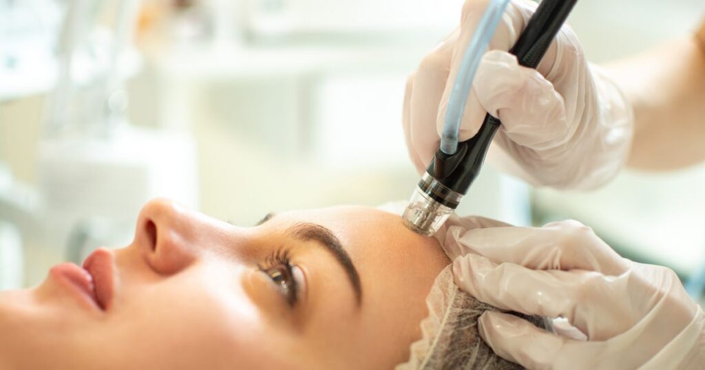 A person receives their first HydraFacial, a skincare treatment using a handheld device. The practitioner, wearing gloves and a head covering, ensures a safe and effective session.