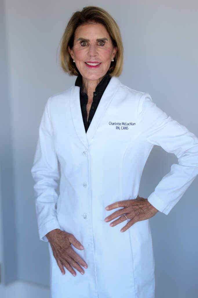 A proud member of Our Team, a woman wearing a white lab coat with "Charlotte McLachlan RN, CANS" embroidered on it stands with one hand on her hip, smiling at the camera.