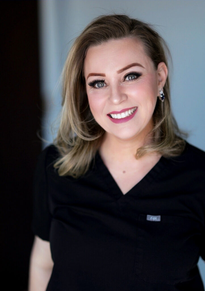 Our Team member with shoulder-length blonde hair, wearing black scrubs, smiles at the camera against a neutral background.