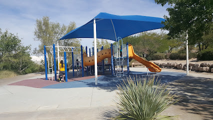 Playground with blue shade cover, slides, climbing equipment, and a child playing; surrounded by trees and desert landscaping under sunny skies.
