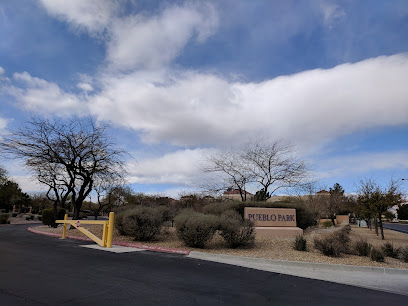 Entrance to Pueblo Park with a yellow gate, bare trees, shrubs, and a partly cloudy sky in the background.