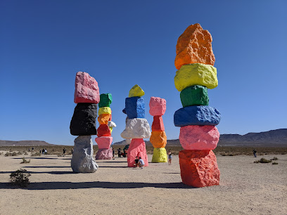 Colorful stacked boulders form tall sculptures in a desert landscape with people nearby under a clear blue sky.