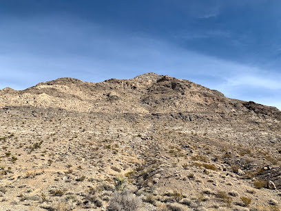 A rocky, barren desert hill under a clear blue sky with sparse vegetation and loose stones scattered across the landscape.