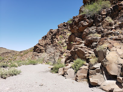 Rocky desert cliff with sparse green shrubs under a clear blue sky, with a dry, sandy path in the foreground.