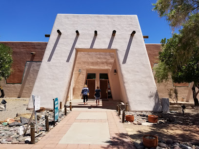 Two people walk toward the entrance of a tan adobe-style building with a distinct geometric archway, surrounded by desert landscaping under a clear blue sky.