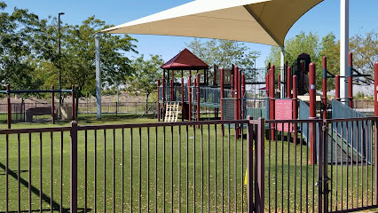 A fenced playground with green artificial turf, climbing structures, slides, and a large shade canopy, surrounded by trees on a sunny day.