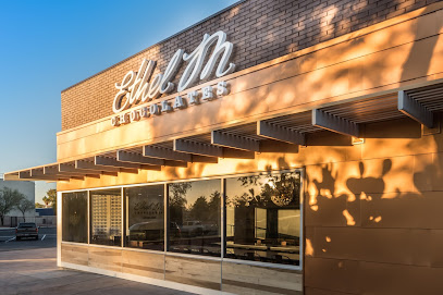 Exterior of Ethel M Chocolates store with brick and tan siding, large windows, and signage reflecting sunlight and tree shadows.