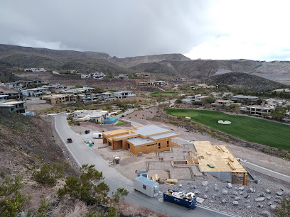 Aerial view of a residential neighborhood under construction near a golf course, with several unfinished houses and construction vehicles visible. Desert hills and cloudy sky in the background.