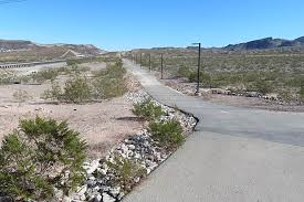 A paved pathway curves through a dry, rocky desert landscape under a clear blue sky, with sparse vegetation and distant hills.
