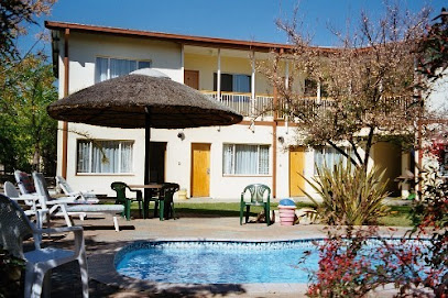 A small outdoor pool surrounded by lounge chairs and a table with green chairs, in front of a two-story building with balconies and trees.