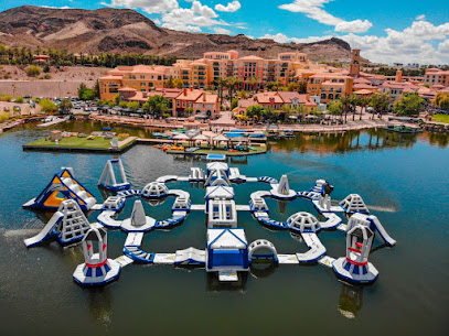 A large inflatable water obstacle course floats on a lake near a resort with buildings and mountains in the background.