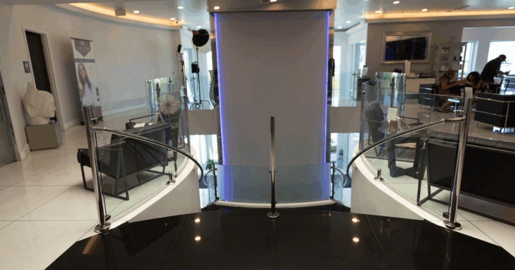 Modern interior space with curved stairs, glass railings, white floors, seating areas, and a vertical light feature in the center—evoking the refined elegance of a body contouring or non-surgical liposuction clinic.