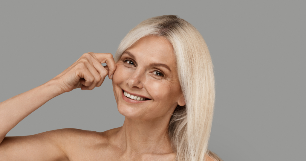 Smiling older woman with long blonde hair pinching her cheek against a plain gray background, suggesting the youthful results achievable with non-surgical lifts and skin tightening treatments in Las Vegas.